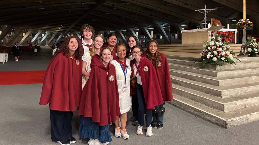 Students in red capes and a faculty member in a white dress stand together and smile in a large chapel Students in red capes and a faculty member in a white dress stand together and smile in a large chapel